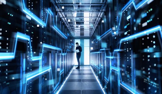 Male IT Engineer Works on a Laptop in a Big Data Center. Rows of Rack Servers are Seen.